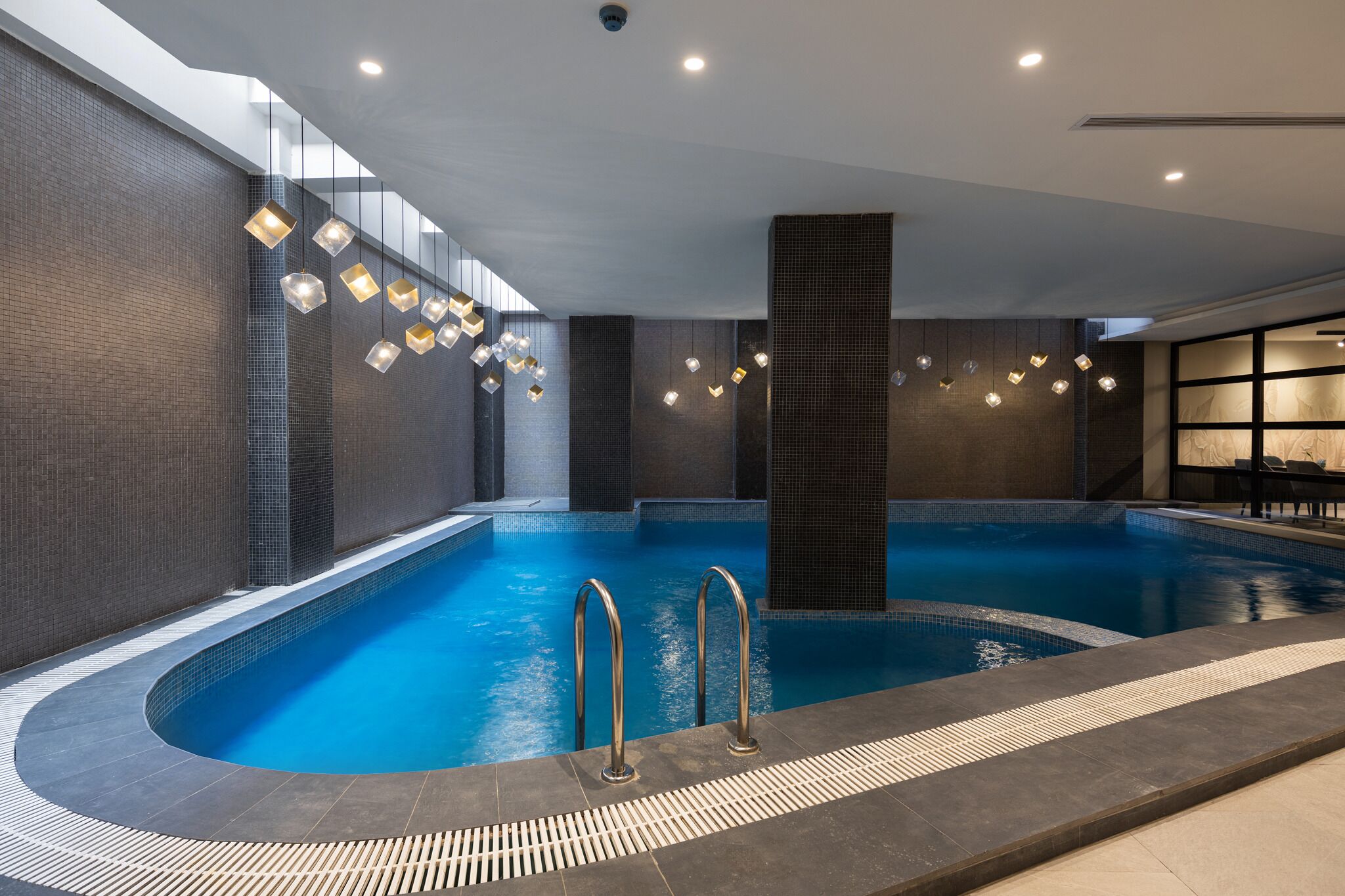 An indoor swimming pool with a tiled edge, a central column, and hanging lights.
