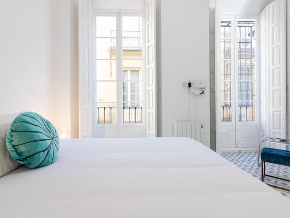 A bedroom with a large bed, a teal pillow, a radiator, and a view of the outside balcony.