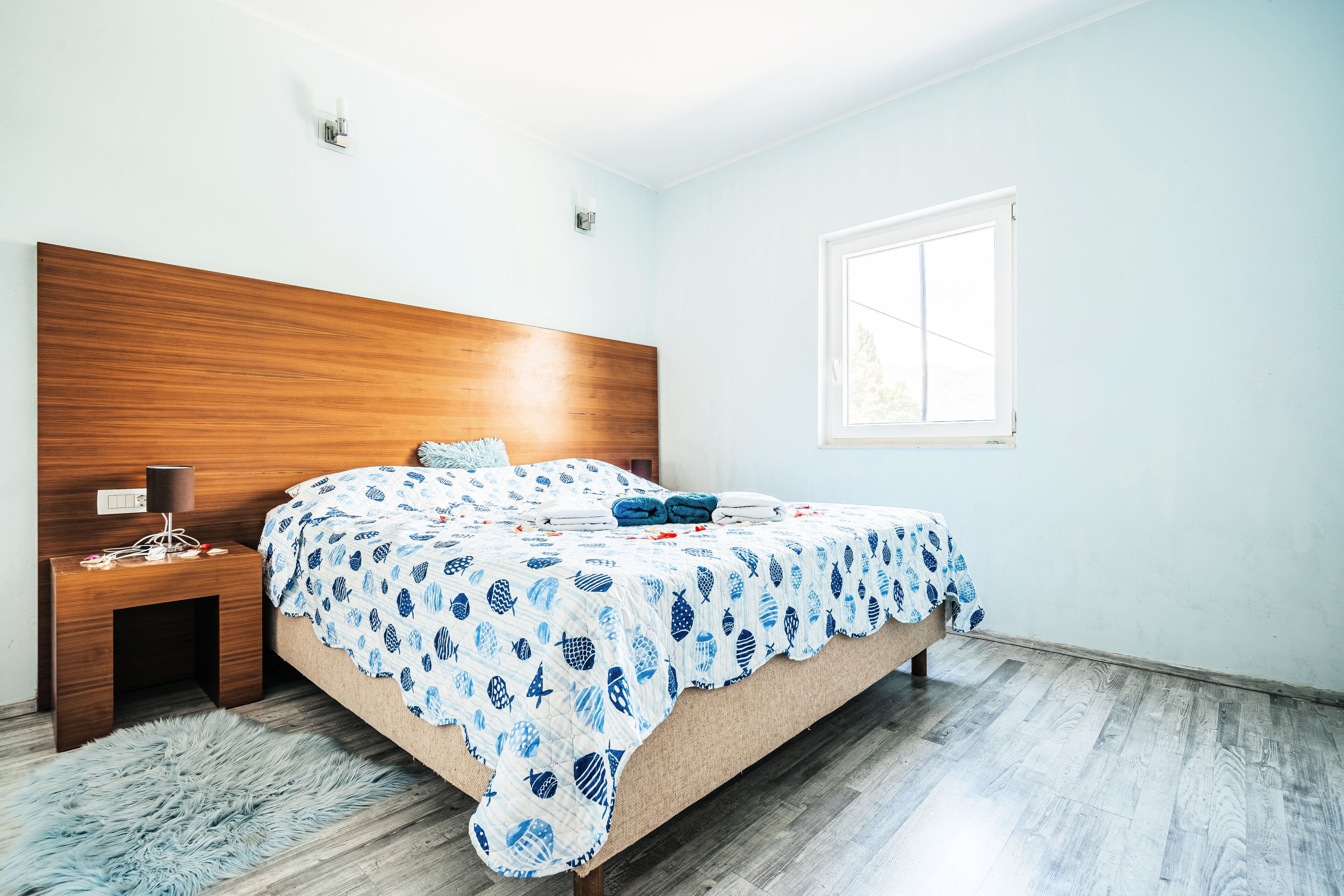 Un dormitorio con cabecera de madera, cama con colcha azul y blanca estampada, mesita de noche con lámpara y ventana que deja entrar luz natural.