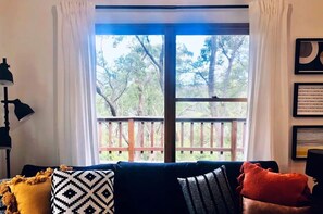 Relax in a serene space with a view of the outdoor deck and trees, featuring a sofa, floor lamp, and personal touches.