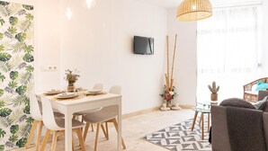 Una sala de estar moderna con comedor, que incluye una mesa blanca, sillas, un televisor montado en la pared y un gran ventanal con luz natural.