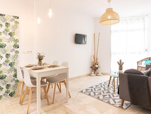 A modern living room with a dining area, featuring a white table, chairs, a TV mounted on the wall, and a large window with natural light.