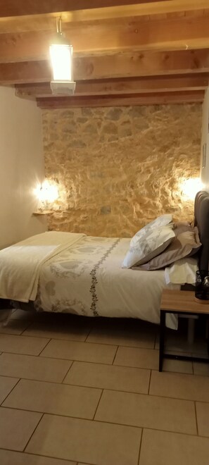 Une chambre avec un mur en pierre, un lit avec une literie à motifs, deux lampes fixées au mur et une table de chevet avec une tasse.