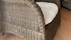 Une chaise en osier avec un coussin, une table en osier et une lampe allumée en arrière-plan.