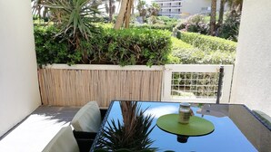 A patio with a glass-top table, white cushioned chairs, a green umbrella, and a view of a landscaped garden with palm trees and a building in the background. - Studio Canet Plage (CANET PLAGE)