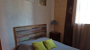 A bedroom with a wooden headboard, a bed with a grey bedspread and two green pillows, a bedside table with a lamp, and a window with curtains. - À 10 Minutes de la Plage, Petite Maison au Calme Avec Patio (Le Barcarès)