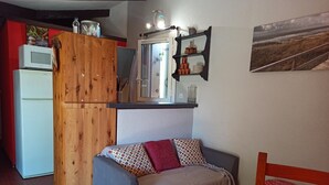 A compact living space with a kitchenette, a wooden cabinet, a sofa with patterned cushions, and a small dining area with red stools. - À 10 Minutes de la Plage, Petite Maison au Calme Avec Patio (Le Barcarès)