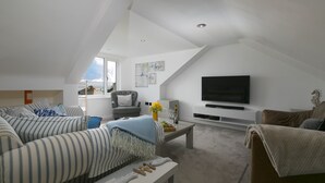 A modern living room with a high ceiling, a flat-screen TV mounted on the wall, a sofa, a coffee table, and a window with a view. - Willows - Sleeps 6 - On-site Parking - Outside Space (St Ives)