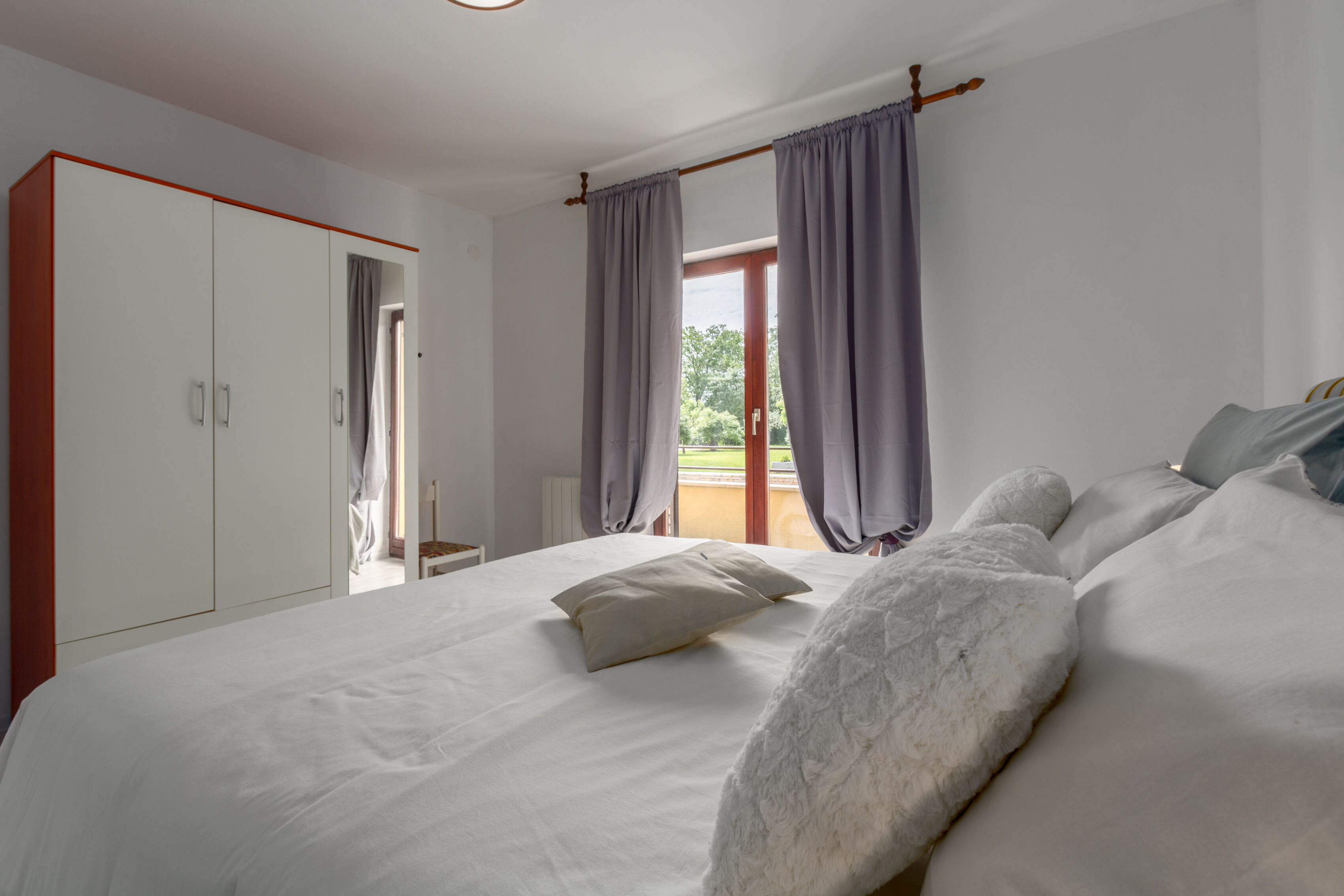 Ein Schlafzimmer mit einem großen Bett, einem Kleiderschrank und Blick ins Freie durch ein Fenster.