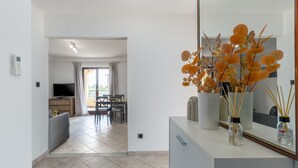 Ein modernes Wohnzimmer mit Sofa, Couchtisch und Fernseher. Es gibt eine Essecke mit Tisch und Stühlen. Auf der Arbeitsplatte steht eine Vase mit orangefarbenen Blumen.
