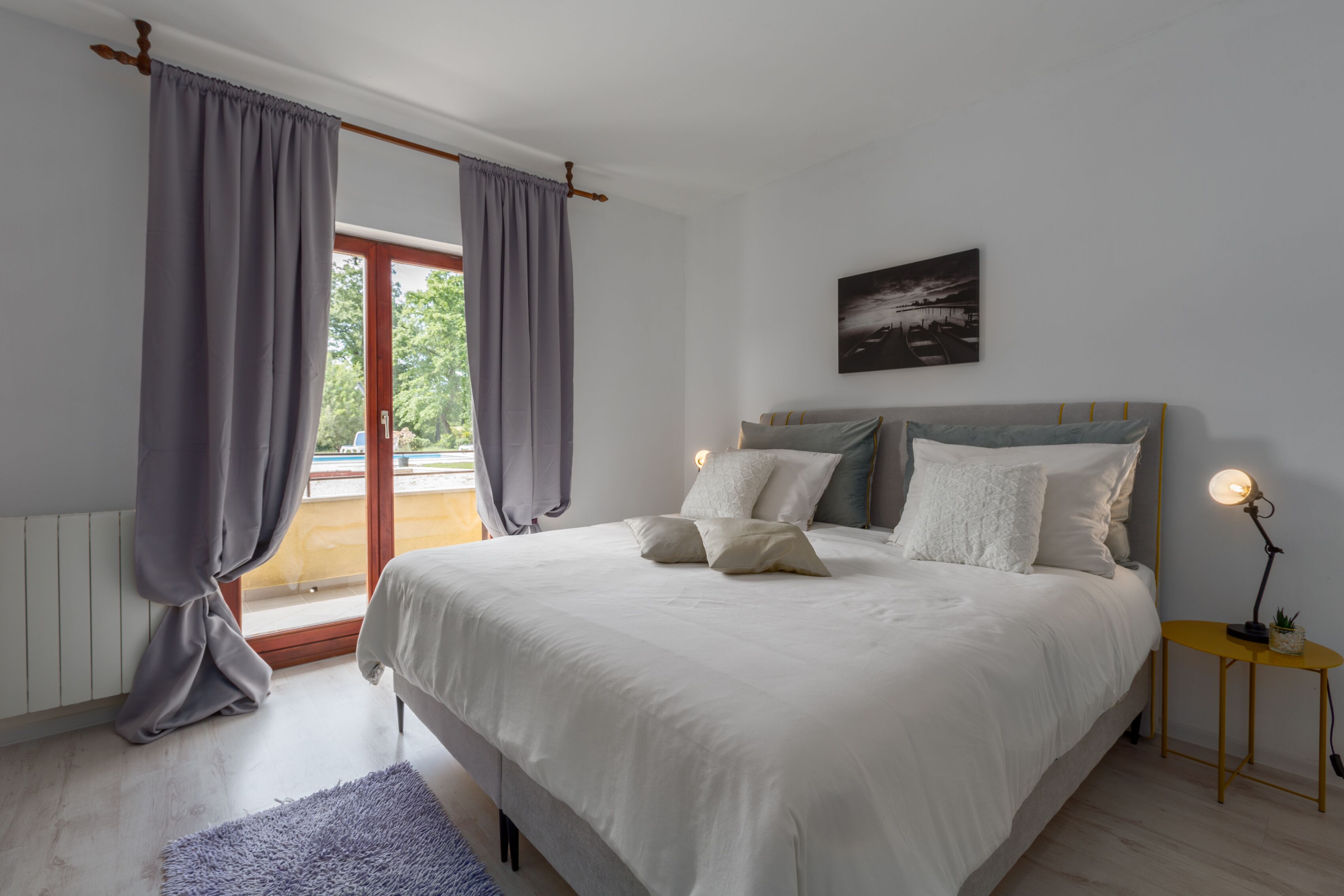 Une chambre à coucher avec un lit, des rideaux, une table de chevet avec une lampe et un tableau accroché au mur.