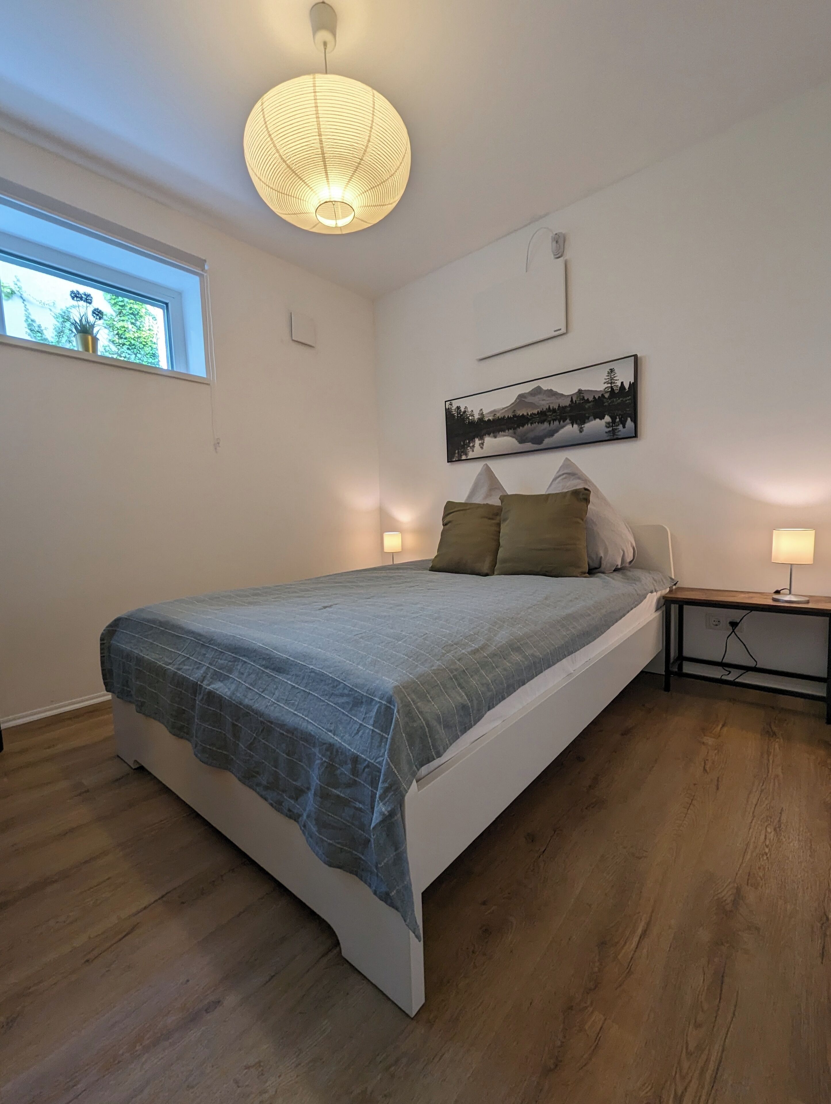 A bedroom with a bed, a window, a wall-mounted picture, and a bedside table with a lamp.