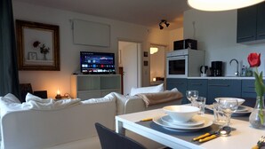 Ein modernes Wohnzimmer mit einem weißen Sofa, einem Esstisch gedeckt mit Tellern, Gläsern und Besteck sowie einer Küchenzeile mit einer Kaffeemaschine und Schränken.