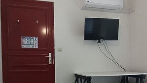 Une pièce avec une porte rouge, un climatiseur blanc, un téléviseur à écran plat fixé au mur, une petite table ronde et deux chaises noires.