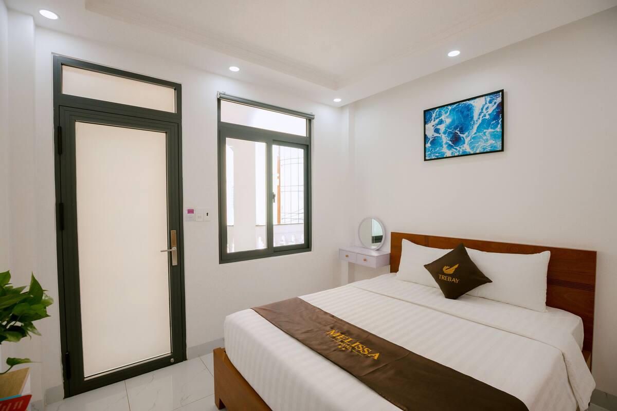 Habitación de hotel con cama, ventana, mesita de noche con espejo y un cuadro en la pared.