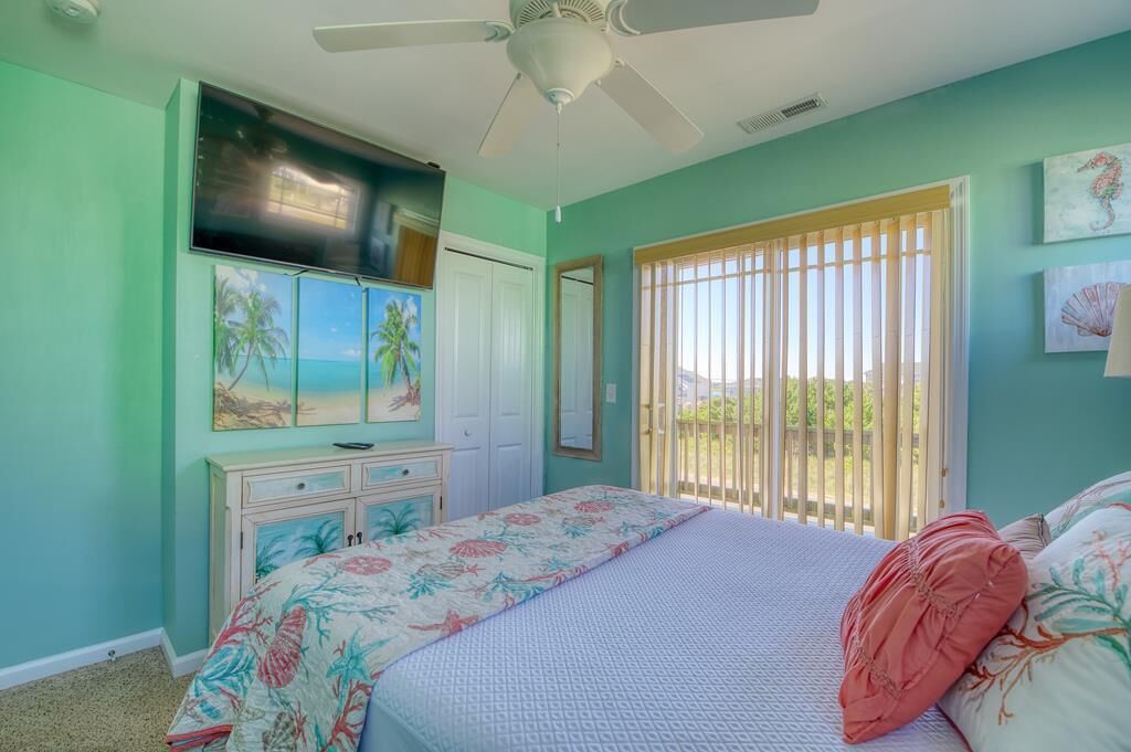 A bedroom with a bed, a ceiling fan, a window with blinds, a dresser, and artwork on the walls.