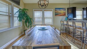 A dining area with a wooden table, chairs, a chandelier, and a large window with blinds. - Your Slice of Heaven on Topsail Island (Topsail Beach)