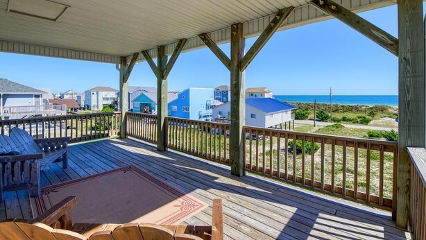 A covered deck with wooden railings, a seating area, and a view of the ocean and beach houses. - Your Slice of Heaven on Topsail Island (Topsail Beach)