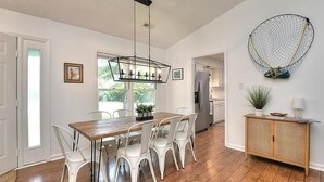 Un comedor con mesa de madera y sillas blancas, un colgante de luz y una consola de mimbre con una planta.