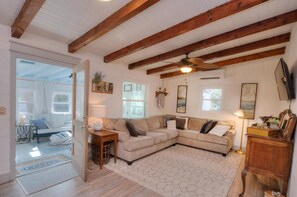 A living room with a beige sectional sofa, wooden beams on the ceiling, a ceiling fan, and a television on the wall.