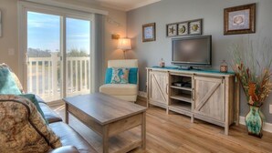 A living room with a sofa, a coffee table, a TV, and a sliding glass door leading to a balcony. - Experience ocean views at the Oak Island OW 9203, a 2-bedroom condo for up to 6 guests. (Oak Island)