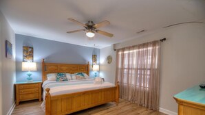 A bedroom with a wooden bed, two bedside tables with lamps, a ceiling fan, a clock on the wall, and a window with curtains. - Experience ocean views at the Oak Island OW 9203, a 2-bedroom condo for up to 6 guests. (Oak Island)