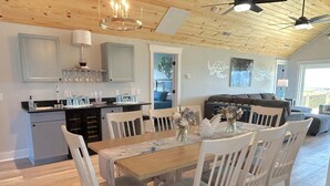 Un comedor con una mesa de madera y sillas blancas, una cocina con armarios grises, y una sala de estar con un sofá y un ventilador de techo.