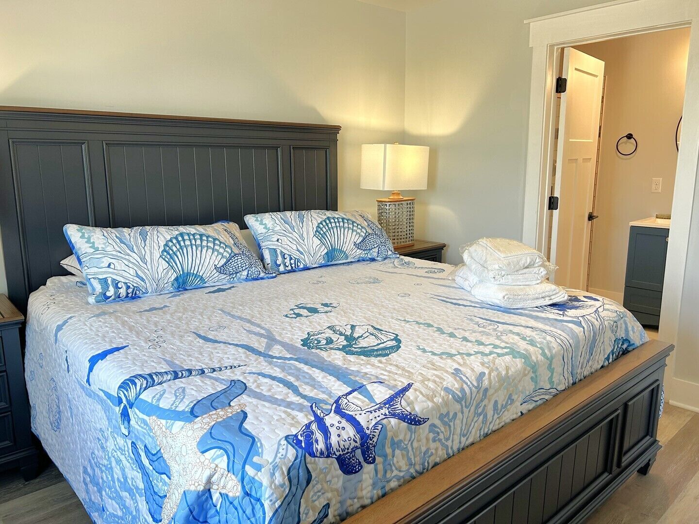 Una cama con colcha temática oceánica en tonos azul y blanco, cabecera de madera y lámpara de noche.