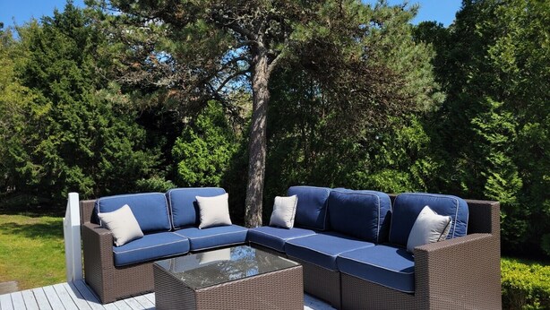 Un sofá seccional exterior con cojines, una mesa de centro de vidrio y un marco de mimbre, ubicado en una terraza de madera rodeada de árboles.