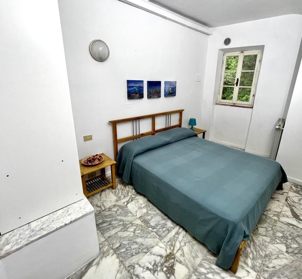 A bedroom with a bed, a nightstand, a small table with a bowl of fruit, and a window with a view of greenery.