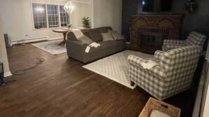 A living room with a sofa, a checkered armchair, a fireplace, and a dining area with a table and chairs. - Highland Hills- Family, Farm, Golf and Goats (Ubly)