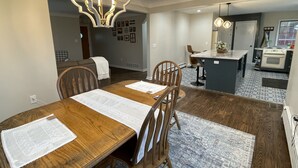 A dining area with a wooden table, chairs, and a chandelier. There is a kitchen area with a stove and sink in the background. - Highland Hills- Family, Farm, Golf and Goats (Ubly)