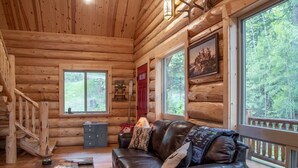 A log cabin interior with a leather sofa, wooden flooring, a fireplace, and large windows offering a view of the forest. - Welcome to Sweet Water, 2 bed 2 bath log cabin 10 minutes from Glacier Park! (Hungry Horse)