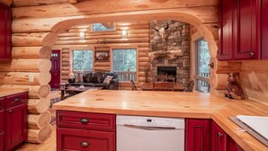 A log cabin interior with a wooden dining area, a stone fireplace, and a kitchen with red cabinets and a dishwasher. - Welcome to Sweet Water, 2 bed 2 bath log cabin 10 minutes from Glacier Park! (Hungry Horse)