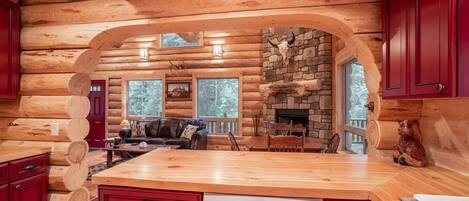 A log cabin interior with a wooden dining area, a stone fireplace, and a kitchen with red cabinets and a dishwasher.