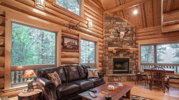 A log cabin interior with a stone fireplace, leather sofa, and large windows overlooking a forest. - Welcome to Sweet Water, 2 bed 2 bath log cabin 10 minutes from Glacier Park! (Hungry Horse)