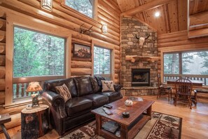 A log cabin interior with a stone fireplace, leather sofa, and large windows overlooking a forest.