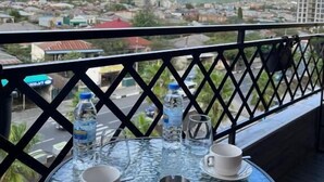 Una mesa redonda de cristal con dos tazas blancas y un posavasos de metal, frente a una barandilla de balcón con vistas a un paisaje urbano con edificios y colinas.