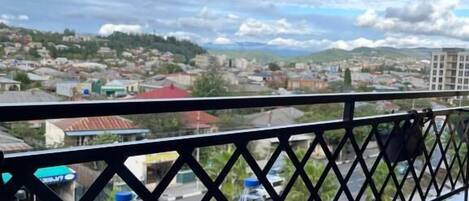 Una mesa redonda de cristal con dos tazas blancas y un posavasos de metal, frente a una barandilla de balcón con vistas a un paisaje urbano con edificios y colinas.
