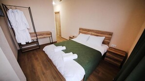 Una cama bien hecha con sábanas blancas y una manta verde, un cabecero de madera, una mesita de noche y un perchero con batas blancas.