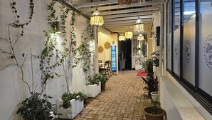 Um corredor estreito com piso de tijolos, plantas em vasos, luminárias pendentes e uma placa.
