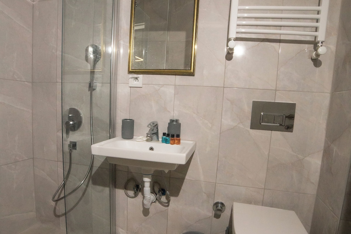 A bathroom with a glass shower enclosure, a white sink with a gold-framed mirror, a toilet, and a towel rack.