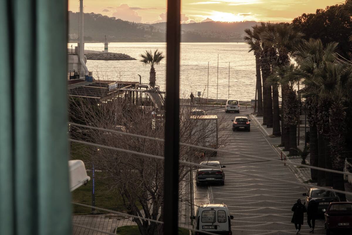 A coastal street with parked cars, a pedestrian, and a view of the sea and a bridge in the distance.