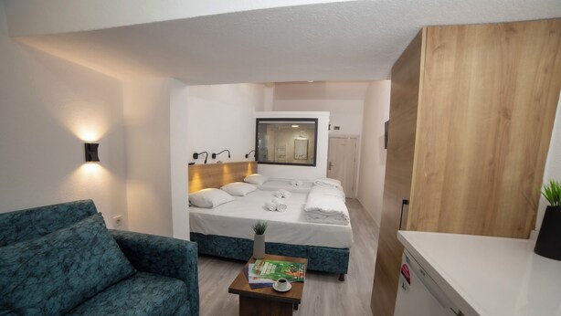 Quarto de hotel pequeno com uma cama, um sofá compacto, uma mesa de cabeceira com um livro e uma xícara, e um armário de madeira.