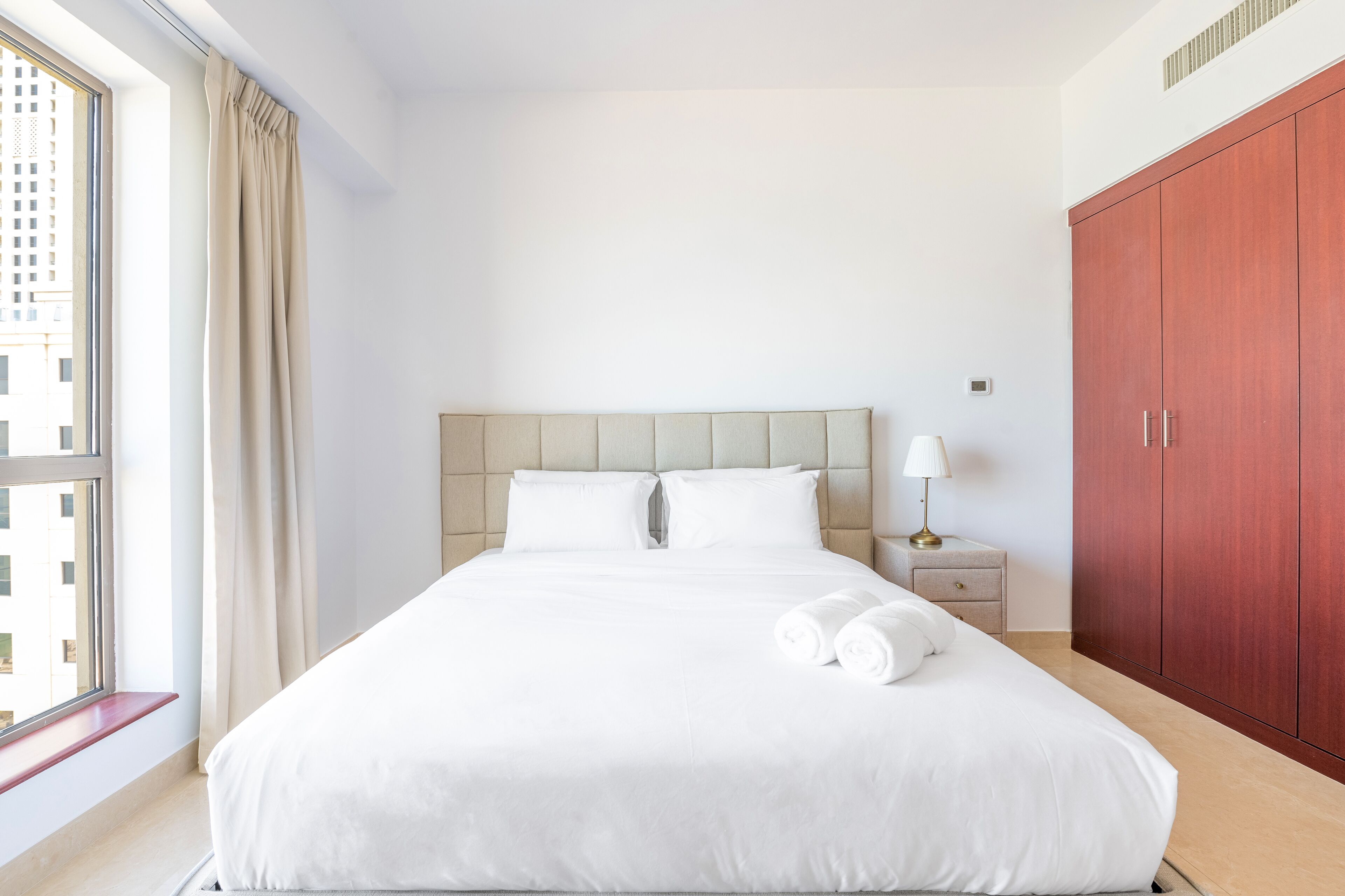 A hotel room with a large bed, a nightstand with a lamp, a wardrobe, and a window with curtains.