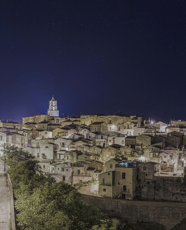 A densely packed hillside town at night with a prominent church tower. - The view of the caves (Grottole)