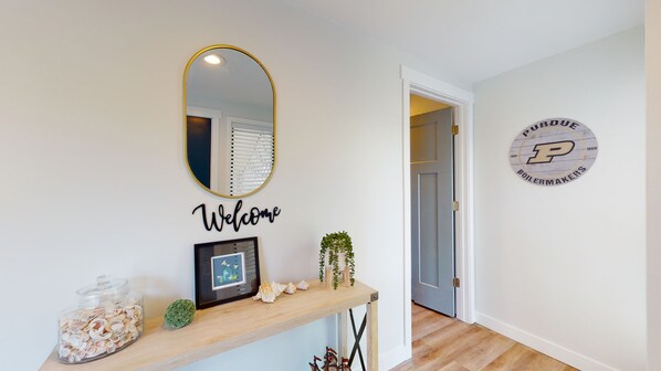 A hallway with a welcome sign, a framed picture, a small plant, and a decorative item on a shelf.