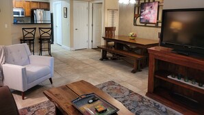 Ein Wohnzimmer mit Fernseher, Couchtisch, Sofa, Esstisch mit Stühlen und einer Küchenzeile im Hintergrund.