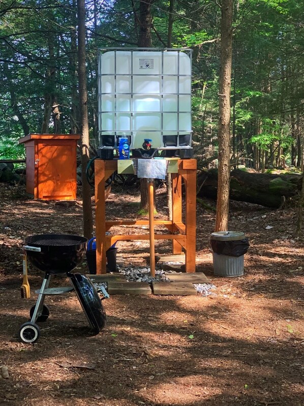 A portable grill setup in a wooded area with a metal container and a small orange cabinet. - Get in touch with nature. (Richmond)