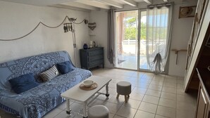 Een woonkamer met een gestreept sofa, een klein tafeltje, twee krukken, een dressoir en een schuifpui naar een buitenruimte.